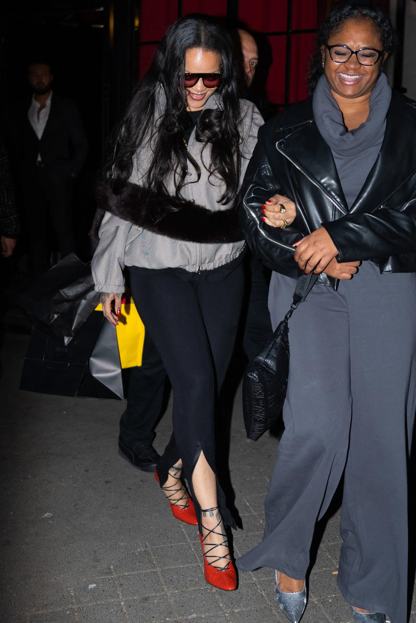 Potret Rihanna di Paris mengenakan jaket bomber abu-abu dengan syal bulu cokelat, legging hitam Balenciaga, dan sepatu hak merah menyala.